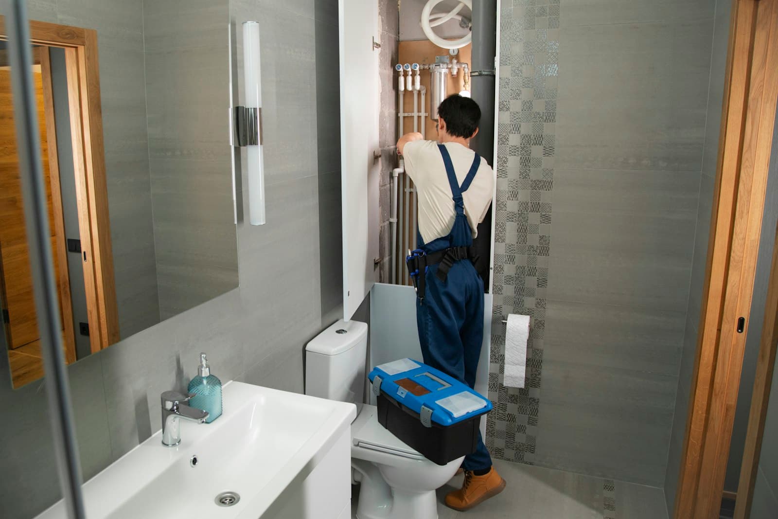 A plumber with a toolbox fixing pipes behind a bathroom wall panel